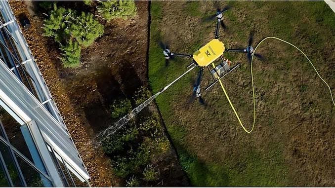 Overhead view of a tethered Drone Precision Solutions cleaning drone soft-washing the glass facade of a building, its supply line running back across the lawn