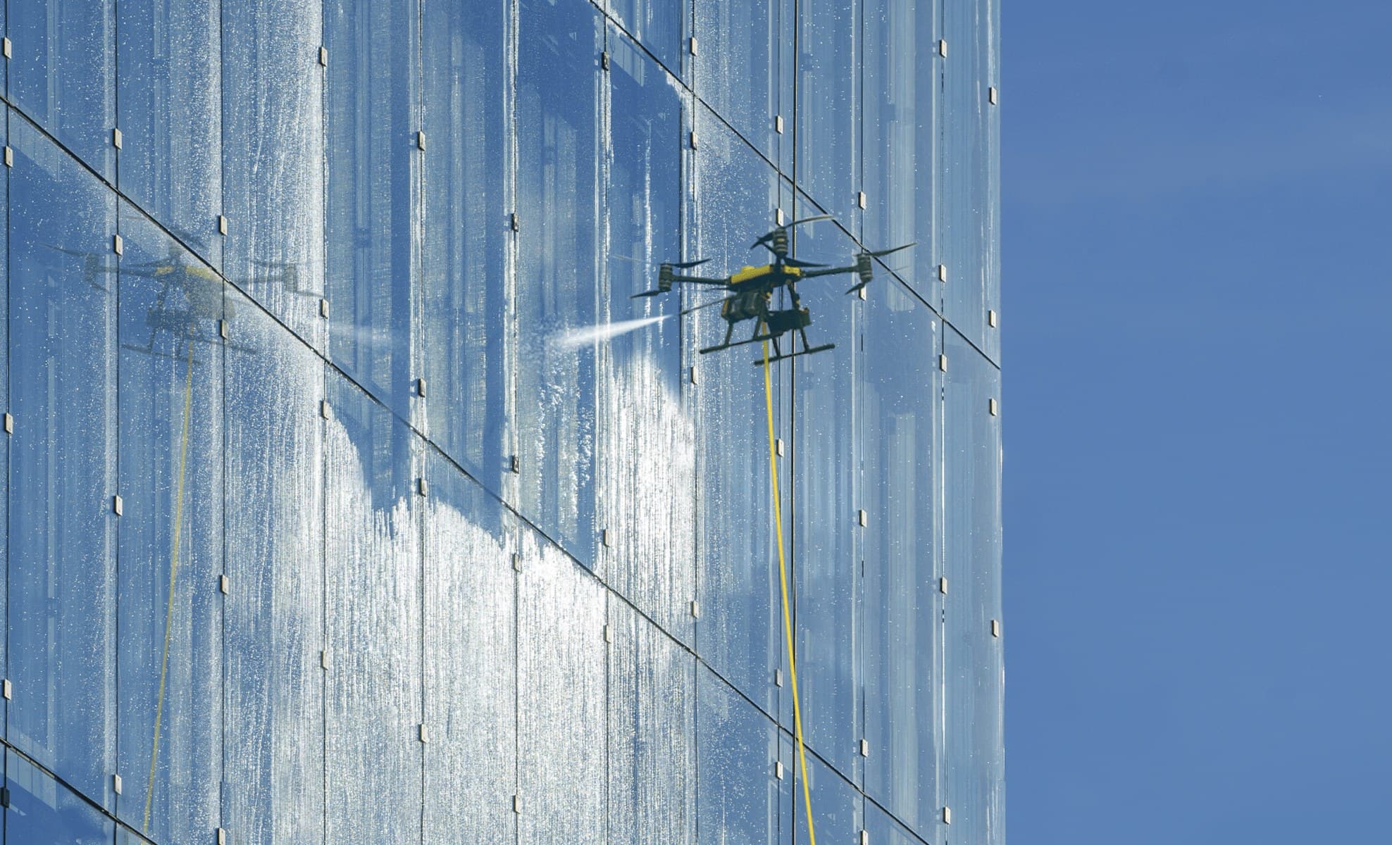 A tethered cleaning drone soft-washing the glass facade of a high-rise building against a clear blue sky