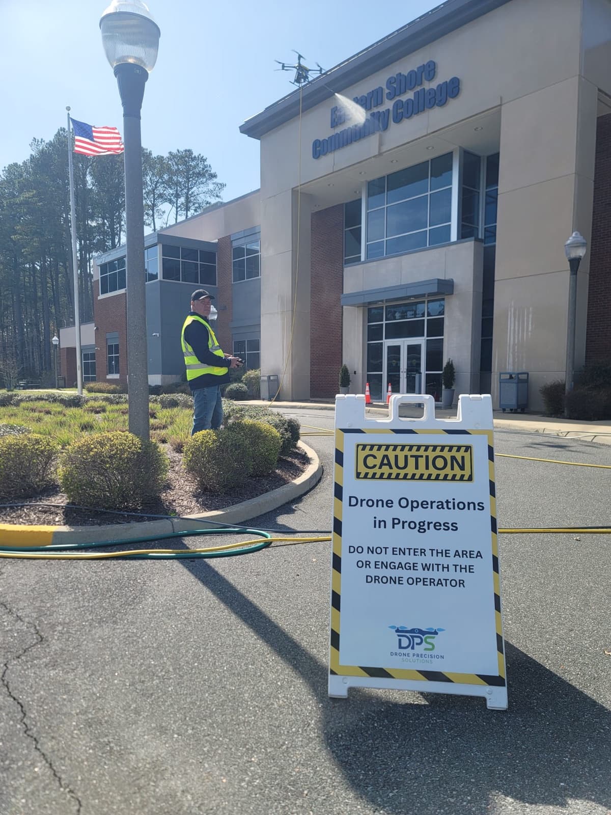 A cleaning drone washing the front of Eastern Shore Community College, with a DPS 'Drone Operations in Progress' caution sign in the foreground.